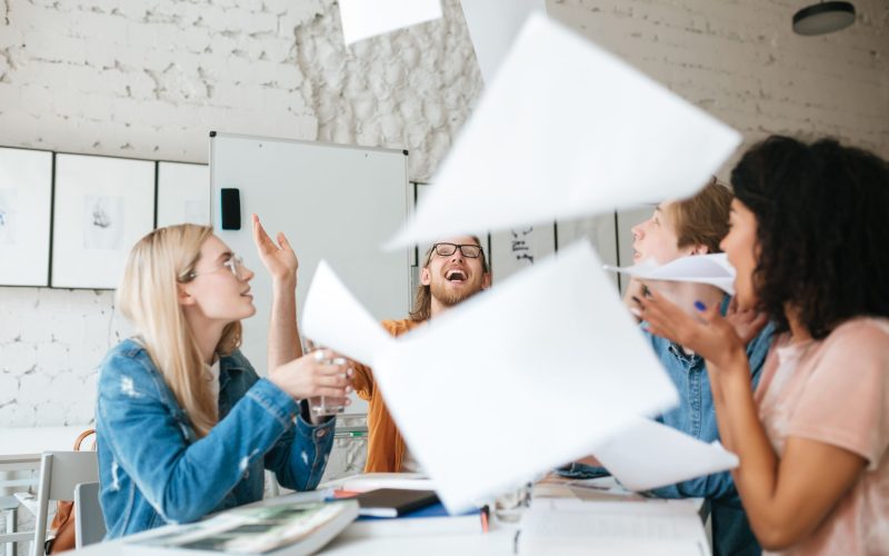 Portrait of emotional people happily throw up papers in office while working together. Group of young students joyfully studying with books on table in classroom.